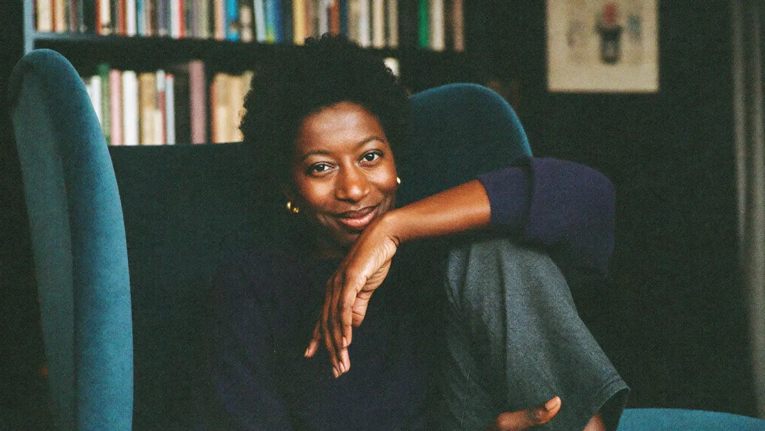 Portrait of a woman resting her arm on the back of an armchair, smiling toward the camera, bookshelves behind her.