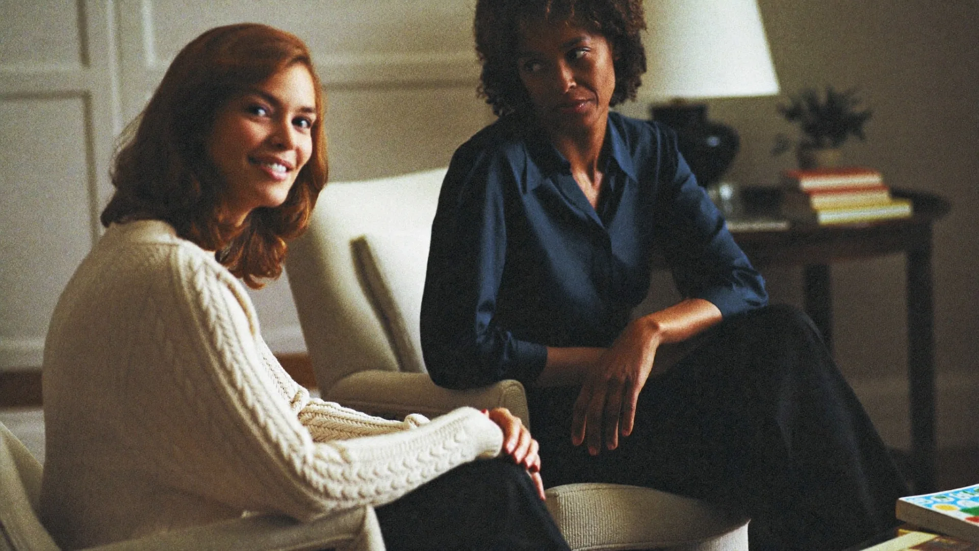 Two women sitting together in a warm front room, mid-conversation, one turned toward the other, listening.